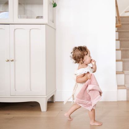  A little girl wearing a white outfit walking through a house holding a Mini Saranoni Lush Blanket that is light pink. The specific color is called Ballet Slipper.
