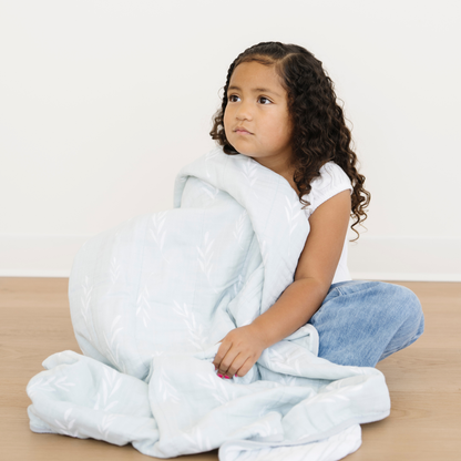 A young girl sits snuggled with an Olive Brand Cotton Muslin -Layer quilt. 