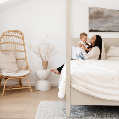 A mother and her son sit on a bed with a Saranoni Grand Faux Fur blanket, featuring an elegant white tone and luxuriously soft texture. This cozy, queen-sized blanket and faux fur blanket offers soothing weight, premium softness, timeless comfort, and is a soft blanket. Perfect as a big blanket and luxury blanket for snuggling or as a luxury bedroom accent, this Saranoni blanket elevates every cozy moment and is a cozy blanket.