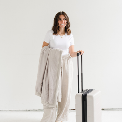 A woman holds a Feather (light gray) Lush XL Saranoni blanket, designed for oversized comfort and featuring a buttery soft, plush texture and has her other hand on a piece of luggage. This luxurious Saranoni blanket is perfect for cozy evenings, reading, or home decor. The Saranoni blanket adds warmth, style, and unmatched softness, making it an ideal gift or essential for ultimate relaxation.