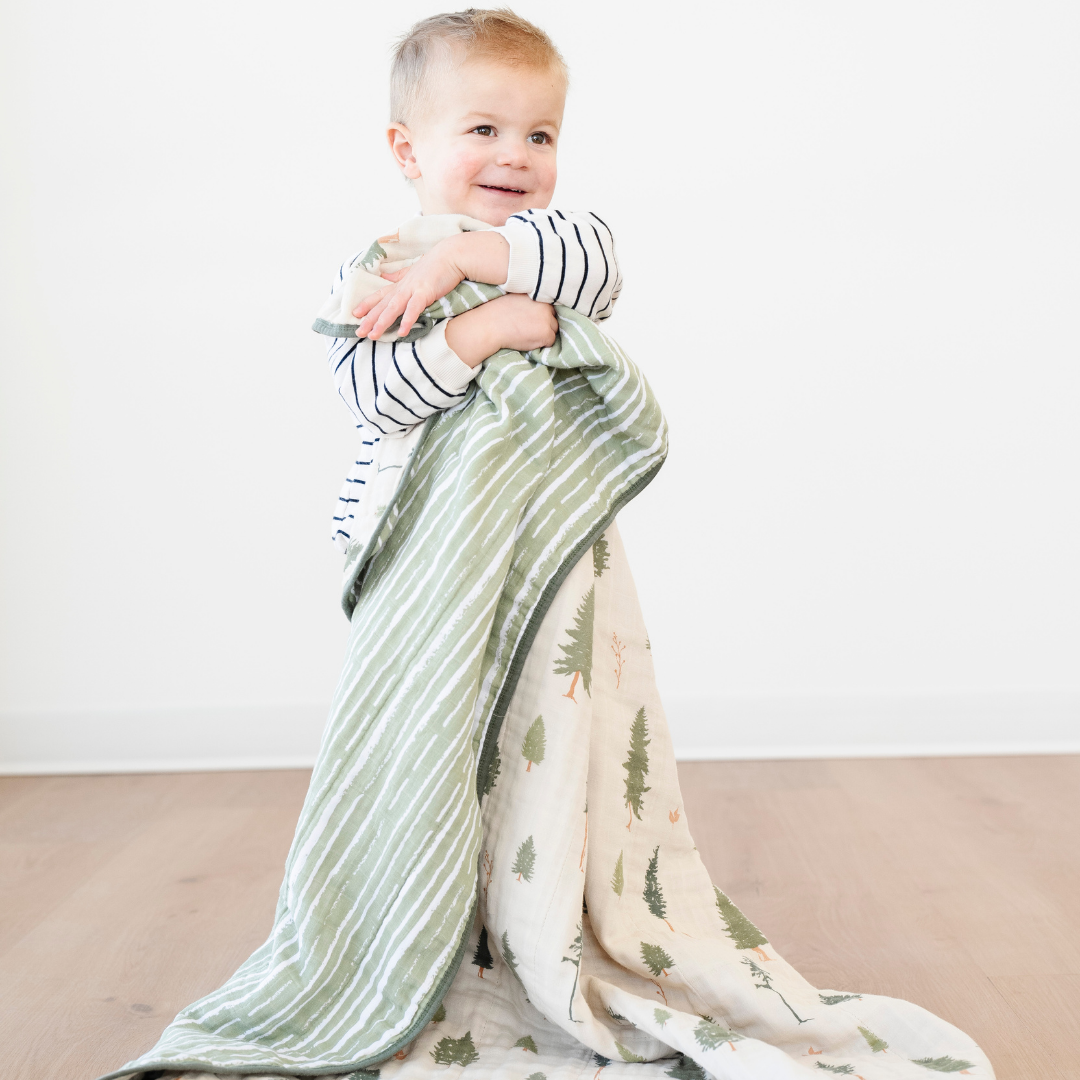A happy little boy is holding a Saranoni blanket that is a cotton muslin quilt. This adorable blanket has pine tree and green striped pattern. This soft blanket is a great toddler gift or baby gift and is so cozy. 