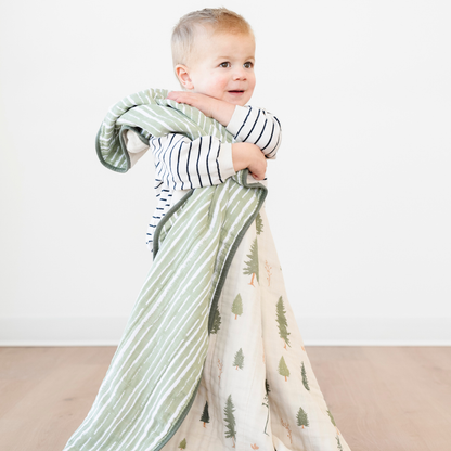 A little boy is hugging a cotton muslin quilt that has a pine tree and green striped pattern on it. This Saranoni blanket is a great toddler blanket or baby blanket. Use as an addition to a baby's nursery or toddler's bedroom. 