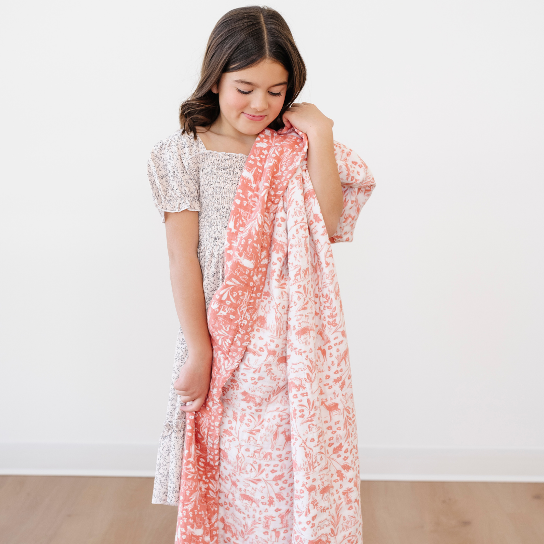 A little girl holds a bamboo rayon muslin quilt that has a woodland pattern on it. This blanket is pink and white. This soft blanket is a Saranoni blanket and is a great baby gift or toddler gift. 
