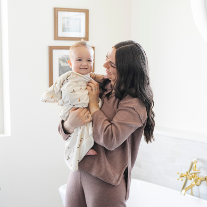 A mother holds her happy baby boy who is wrapped in a Saranoni luxury hooded towel, featuring a soft fabric for quick-drying comfort. This plush baby towel is perfect for bath time, pool time, or beach days, offering a cozy hood and gentle fabric designed for newborns, infants, and toddlers. Adorable baby towel. A must-have baby bath essential, baby shower gift, and new mom must-have. The hooded towel has a print with people on it. The ultimate newborn hooded towel, kids towel, and baby bath towel. 