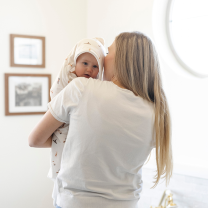 A mother holds her baby girl who is wrapped in a Saranoni luxury hooded towel, featuring a soft fabric for quick-drying comfort. This plush baby towel is perfect for bath time, pool time, or beach days, offering a cozy hood and gentle fabric designed for newborns, infants, and toddlers. Adorable baby towel. A must-have baby bath essential, baby shower gift, and new mom must-have. The hooded towel has a print with people on it. The ultimate newborn hooded towel, kids towel, and baby bath towel. 