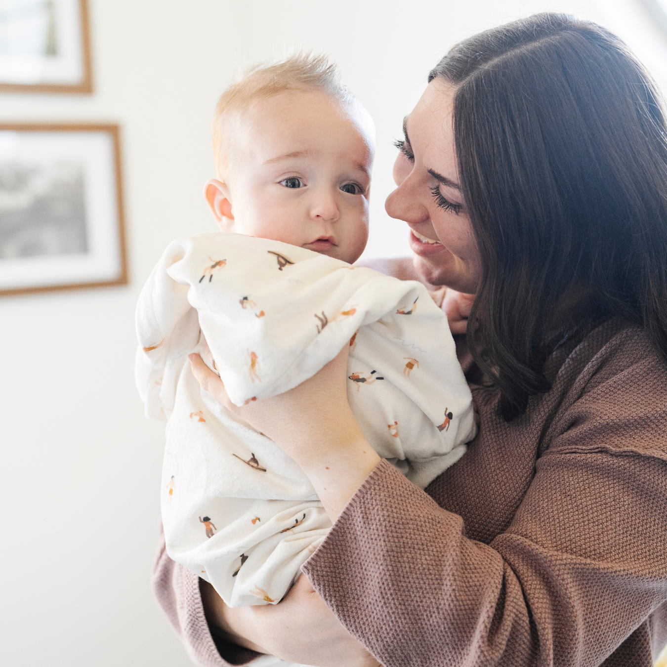 A mother holds her happy baby boy who is wrapped in a Saranoni luxury hooded towel, featuring a soft fabric for quick-drying comfort. This baby towel is perfect for bath time, pool time, or beach days, offering a cozy hood and fabric designed for newborns, infants, and toddlers. Adorable baby towel. A must-have baby bath essential, baby shower gift, and new mom must-have. The hooded towel has a print with people on it. The ultimate newborn hooded towel, kids towel, newborn towel, and baby bath towel. 