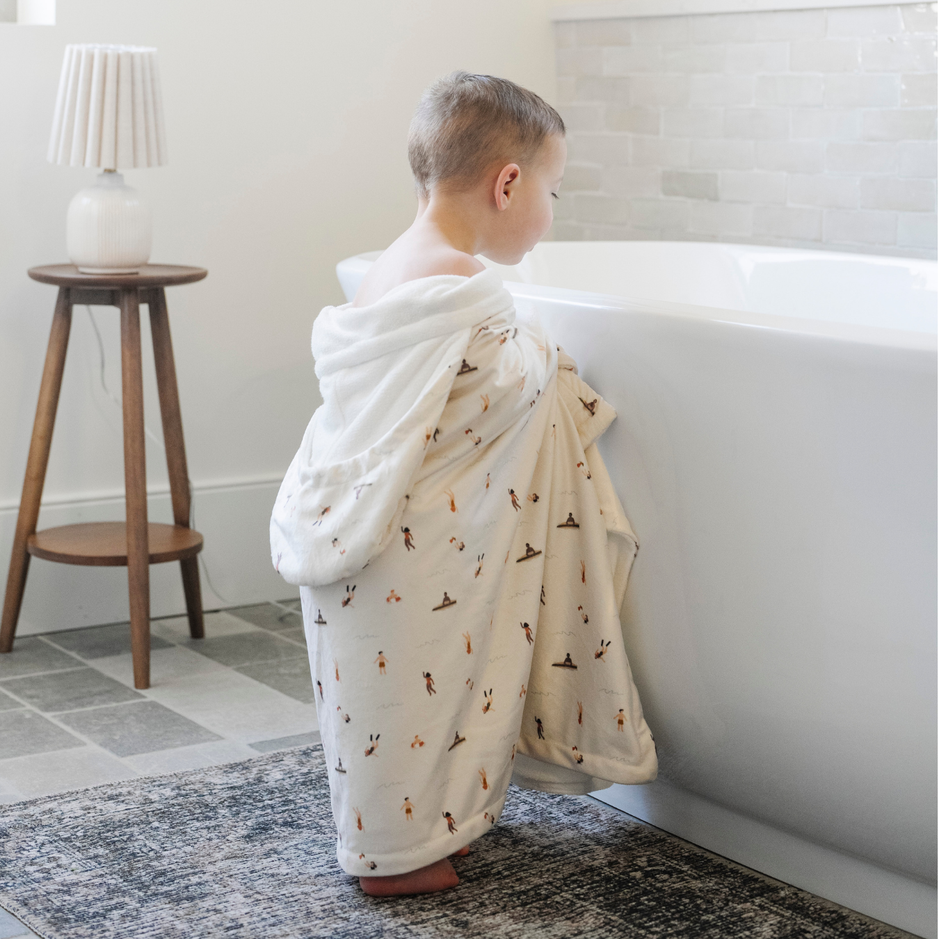A little boy is wrapped in a Saranoni hooded towel and looking into a bathtub. This luxury hooded towel is perfect for bath time, pool days, and beach outings, offering quick-drying warmth while being gentle on sensitive skin. Designed as a toddler hooded towel, it provides a cozy, plush feel that keeps little ones warm after water play. Ideal for parents looking for the best soft toddler towel that combines style, durability, and ultimate comfort. This kid's towel has a print with people on it.