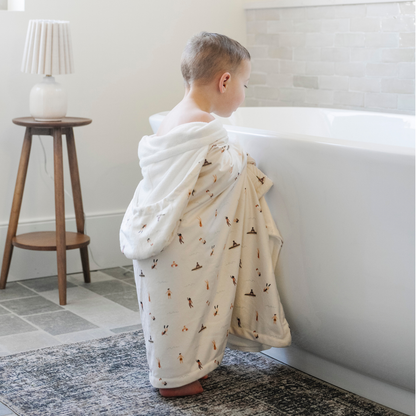 A little boy is wrapped in a Saranoni hooded towel and looking into a bathtub. This luxury hooded towel is perfect for bath time, pool days, and beach outings, offering quick-drying warmth while being gentle on sensitive skin. Designed as a toddler hooded towel, it provides a cozy, plush feel that keeps little ones warm after water play. Ideal for parents looking for the best soft toddler towel that combines style, durability, and ultimate comfort. This kid's towel has a print with people on it.