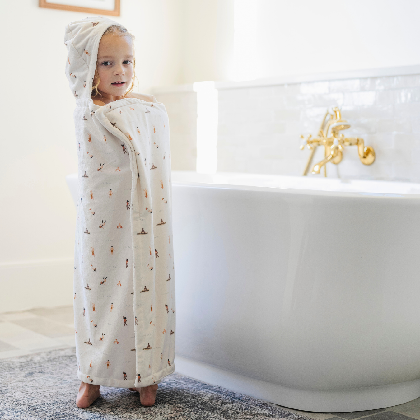 A little girl is wrapped in a Saranoni hooded towel. This luxury hooded towel is perfect for bath time, pool days, and beach outings, offering quick-drying warmth while being gentle on sensitive skin. Designed as a toddler hooded towel, it provides a cozy, plush feel that keeps little ones warm after water play. Ideal for parents looking for the best soft toddler towel that combines style, durability, and ultimate comfort. This kid's towel has a print with people on it. This is a hooded towel for toddlers. 