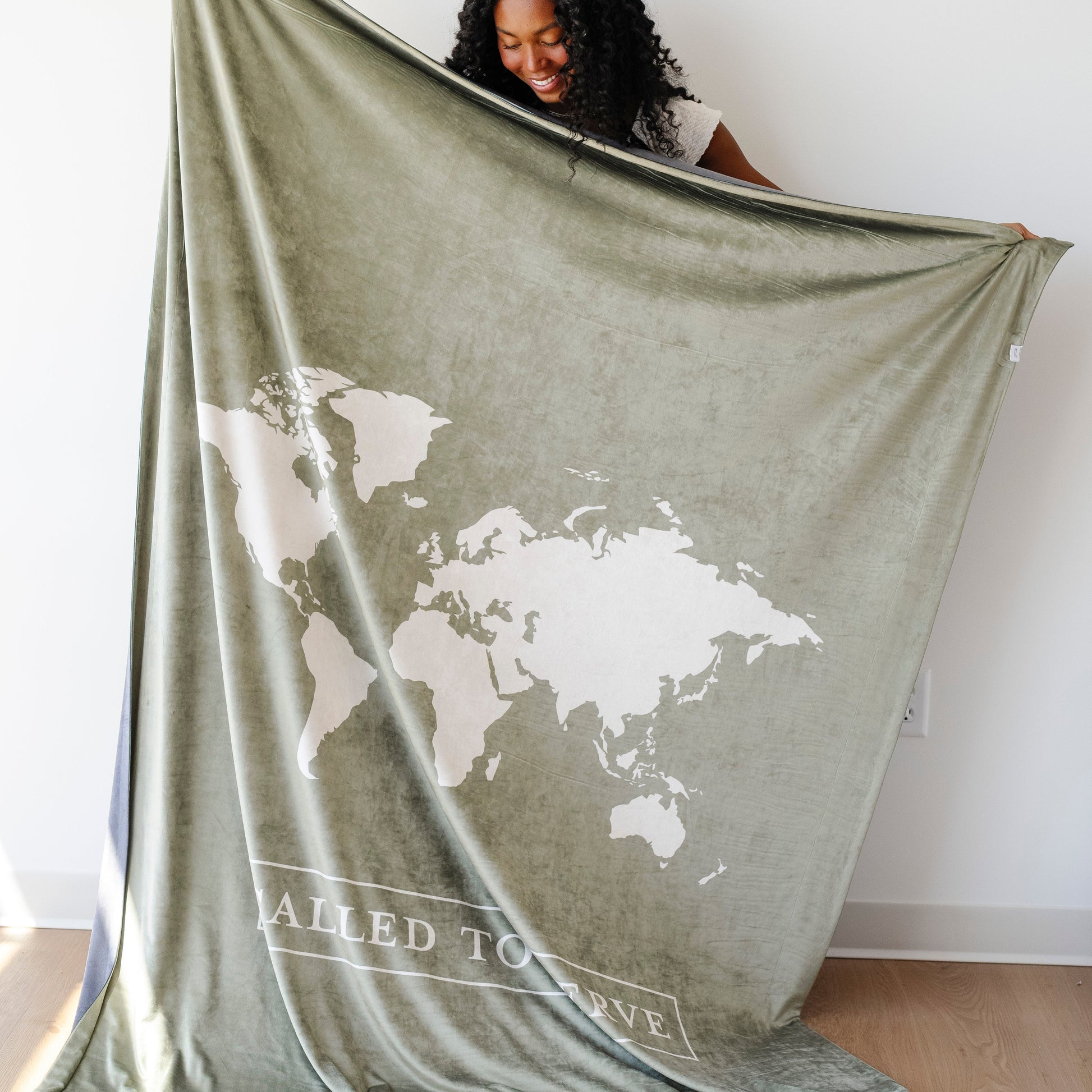 Woman holding a cypress green LDS missionary blanket featuring a world map design and the words "Called to Serve," meaningful missionary gift blanket celebrating faith, service, and missionary journeys around the world.