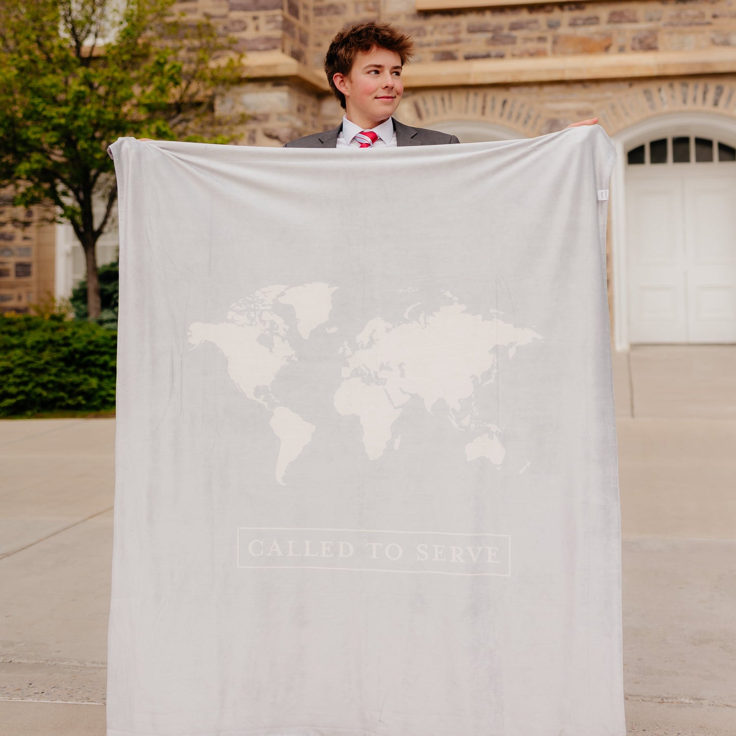 Young missionary holding a shell cream LDS missionary blanket featuring a world map design and the words "Called to Serve," meaningful missionary gift blanket celebrating faith, service, and missionary journeys around the world.