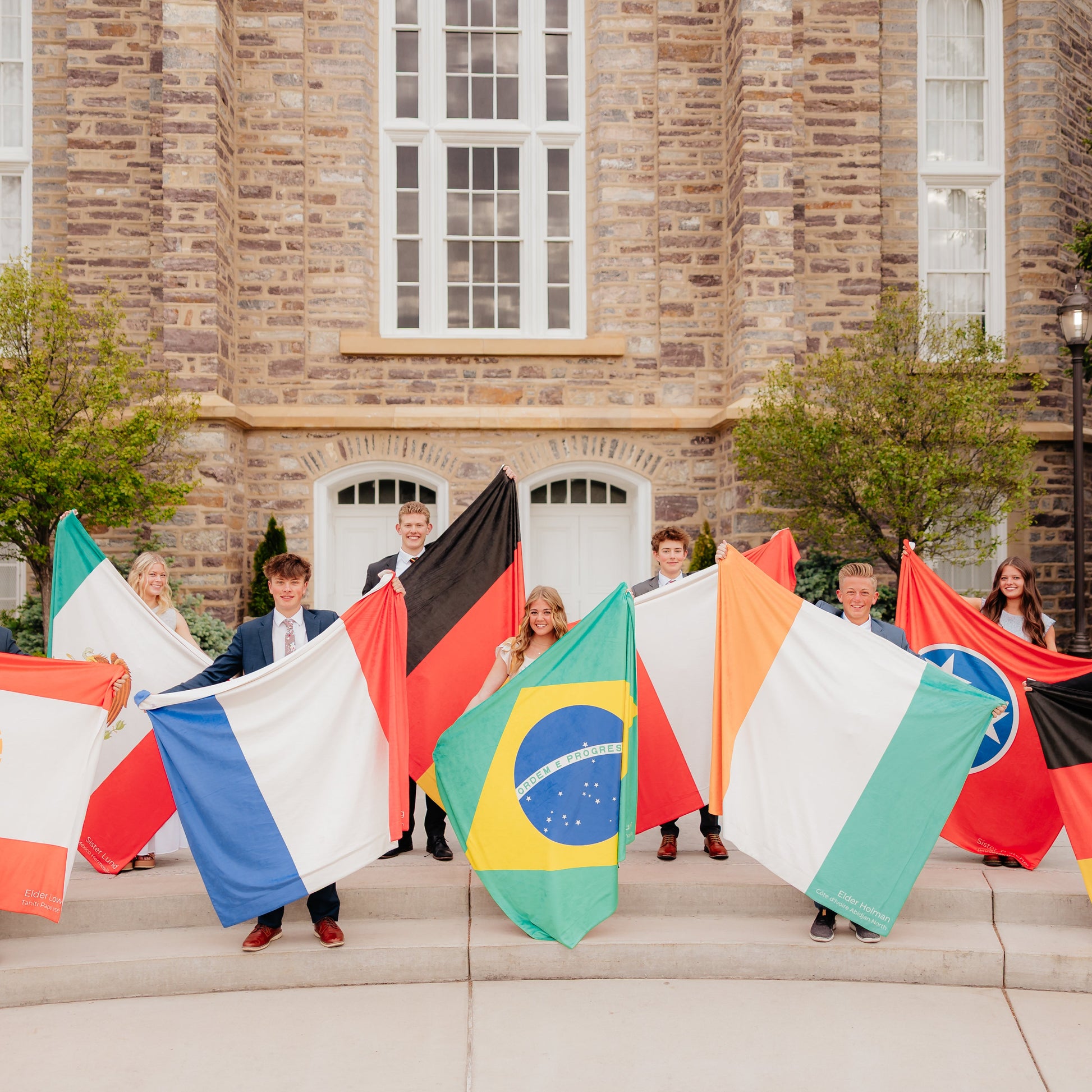LDS missionaries holding personalized mission flag blankets in front of a temple, featuring custom country flags and missionary service locations.