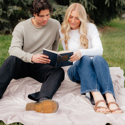 Man and woman reading together on a tan Fishers of Men Christian blanket outdoors, a thoughtful Christian gift