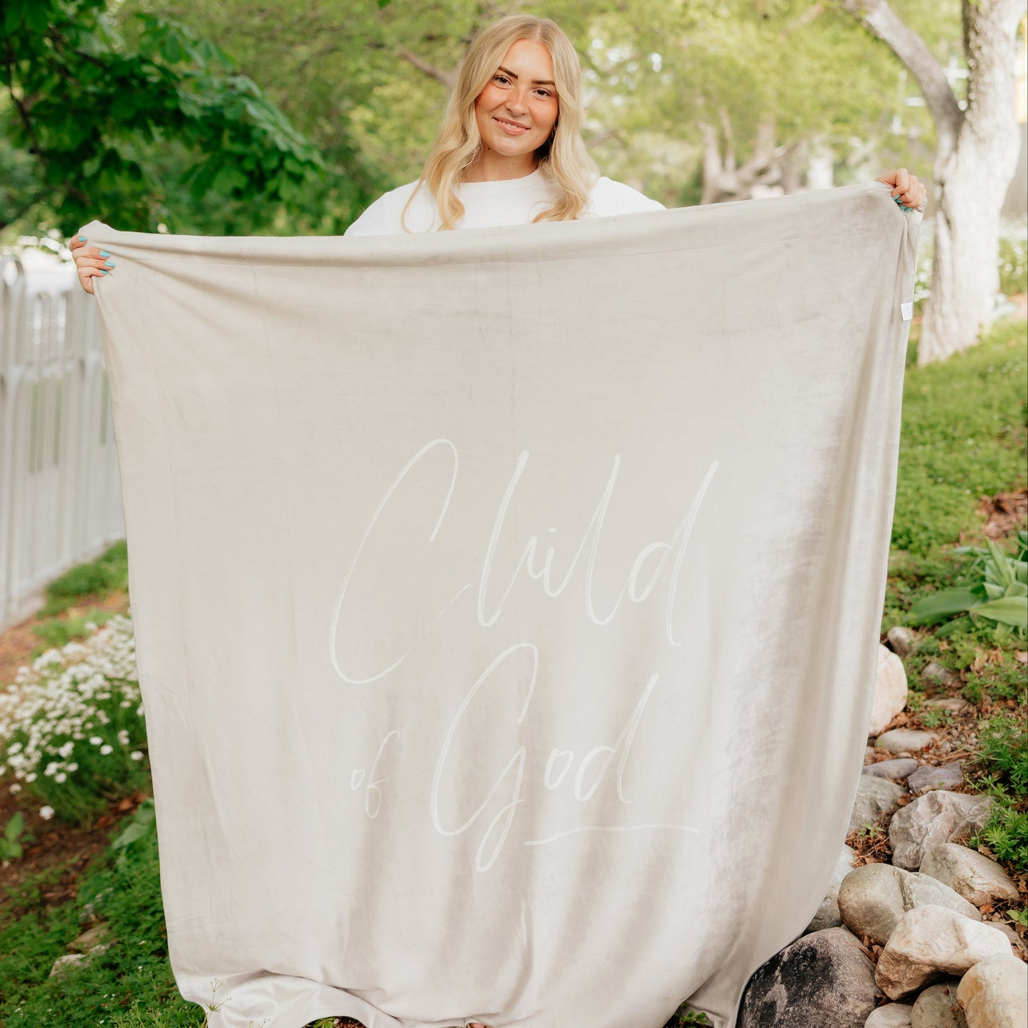 Woman holding a soft taupe Christian blanket featuring the words "Child of God" in elegant cursive lettering.