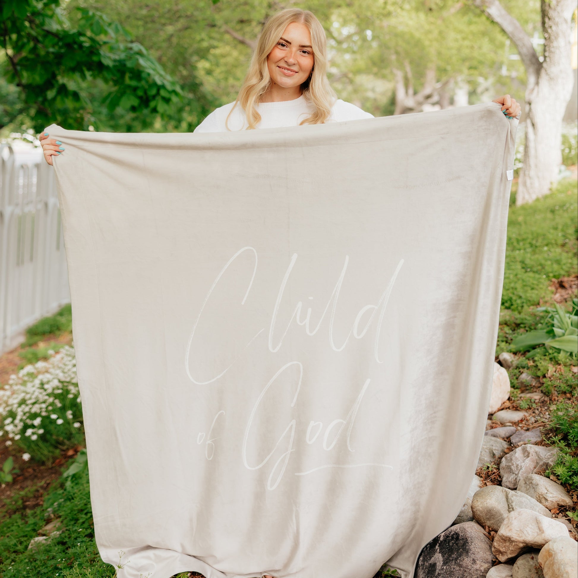 Woman holding a soft taupe Christian blanket featuring the words "Child of God" in elegant cursive lettering.