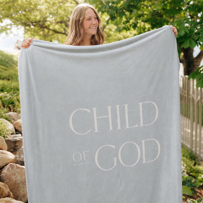 Person holding a light glacier blue Christian blanket featuring the words "Child of God" outdoors, soft faith-inspired throw blanket for comfort, encouragement, and reflection.
