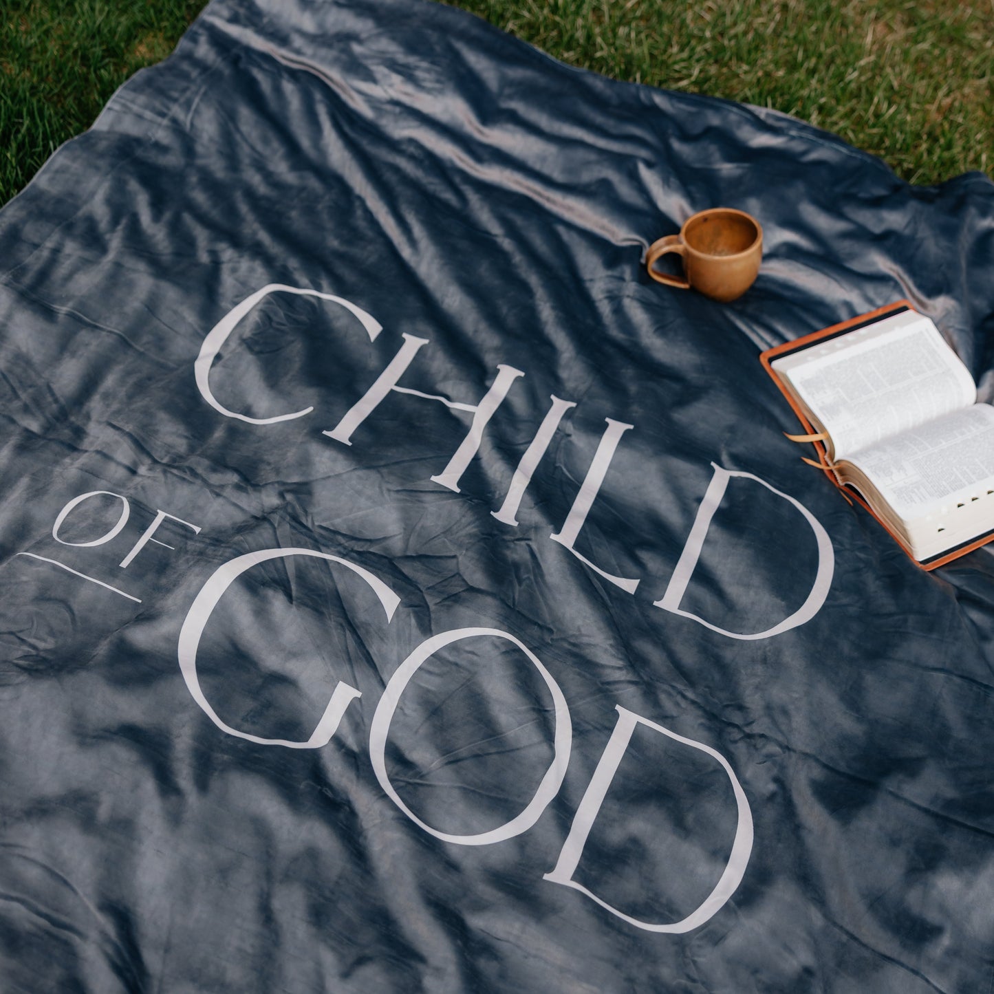 Dark obsidian Christian blanket featuring the words "Child of God" displayed outdoors with an open Bible and mug, faith-inspired throw blanket for comfort and reflection.