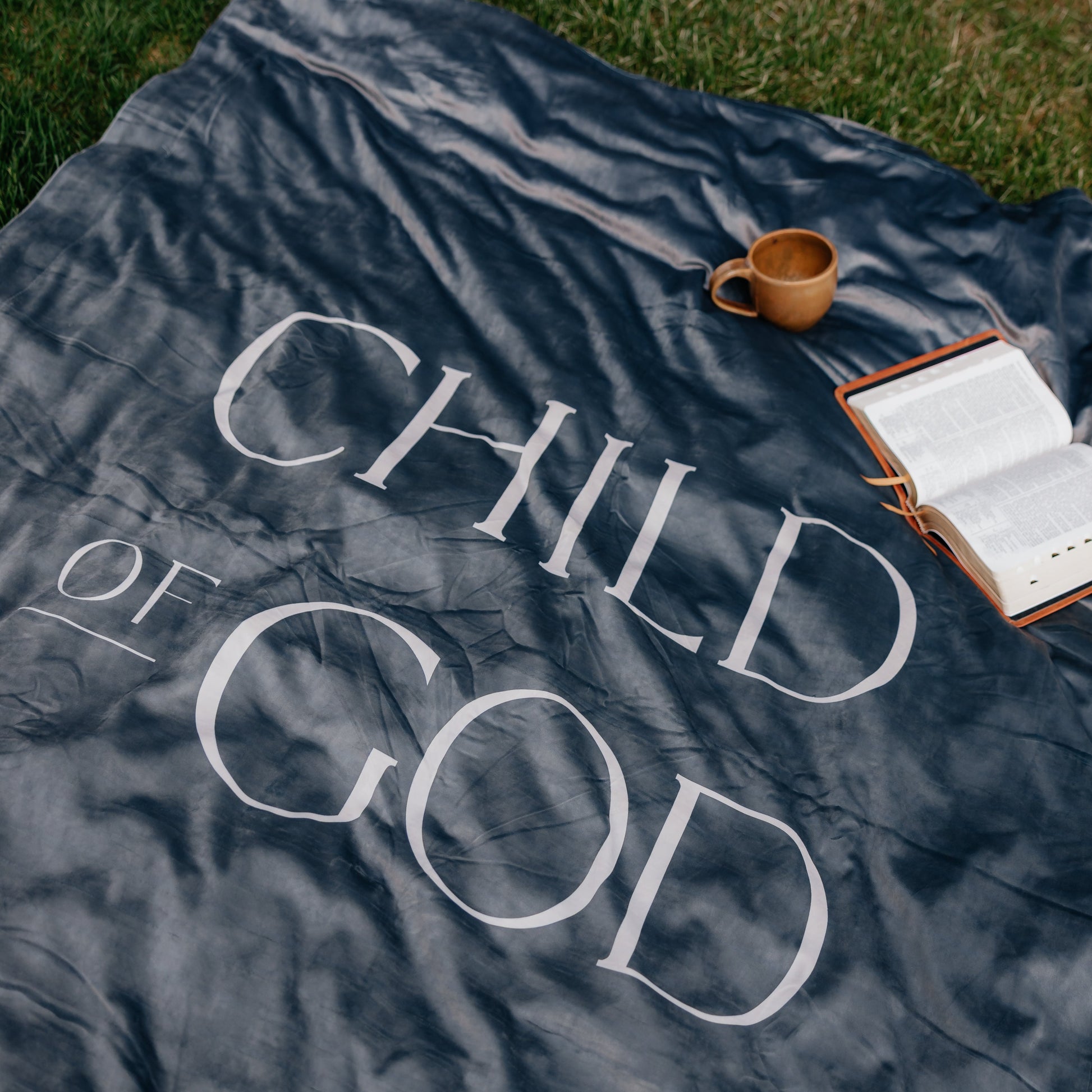 Dark obsidian Christian blanket featuring the words "Child of God" displayed outdoors with an open Bible and mug, faith-inspired throw blanket for comfort and reflection.