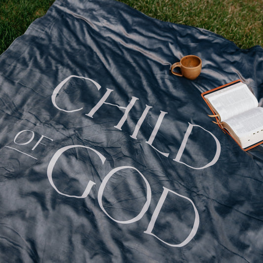 Dark obsidian Christian blanket featuring the words "Child of God" displayed outdoors with an open Bible and mug, faith-inspired throw blanket for comfort and reflection.