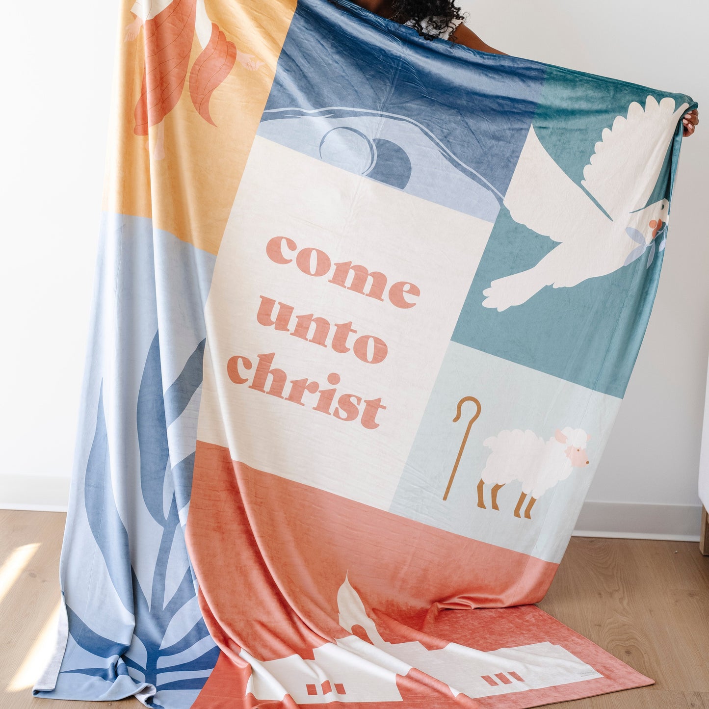 Person holding a Come Unto Christ LDS blanket featuring faith-centered illustrations of Jesus Christ, a dove, shepherd imagery, and uplifting Christian artwork, meaningful blanket for LDS youth and missionaries.