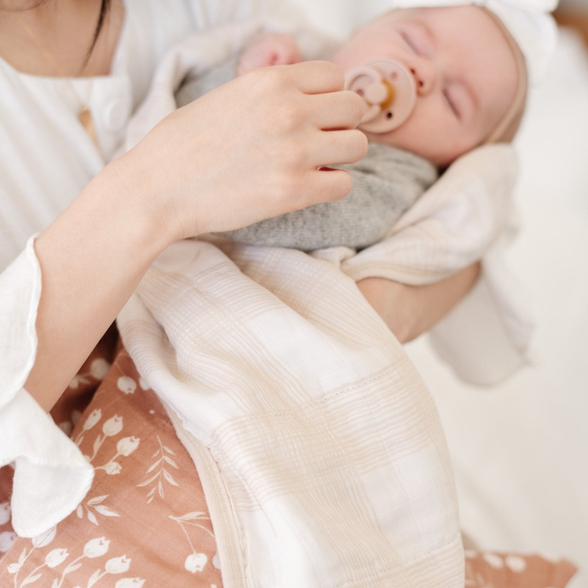 A mother holds her daughter in a Saranoni blanket. The baby is wrapped in a beautifully soft and breathable muslin quilt, designed for both comfort and style. The quilt features a warm, earthy rust tone with delicate white botanical accents, adding a timeless and elegant touch. Its airy, multi-layered muslin fabric is crafted to become even softer with each wash, making it a perfect choice for snuggles, naps, and everyday moments. The soft blanket is a luxury blanket and a Saranoni blanket. 