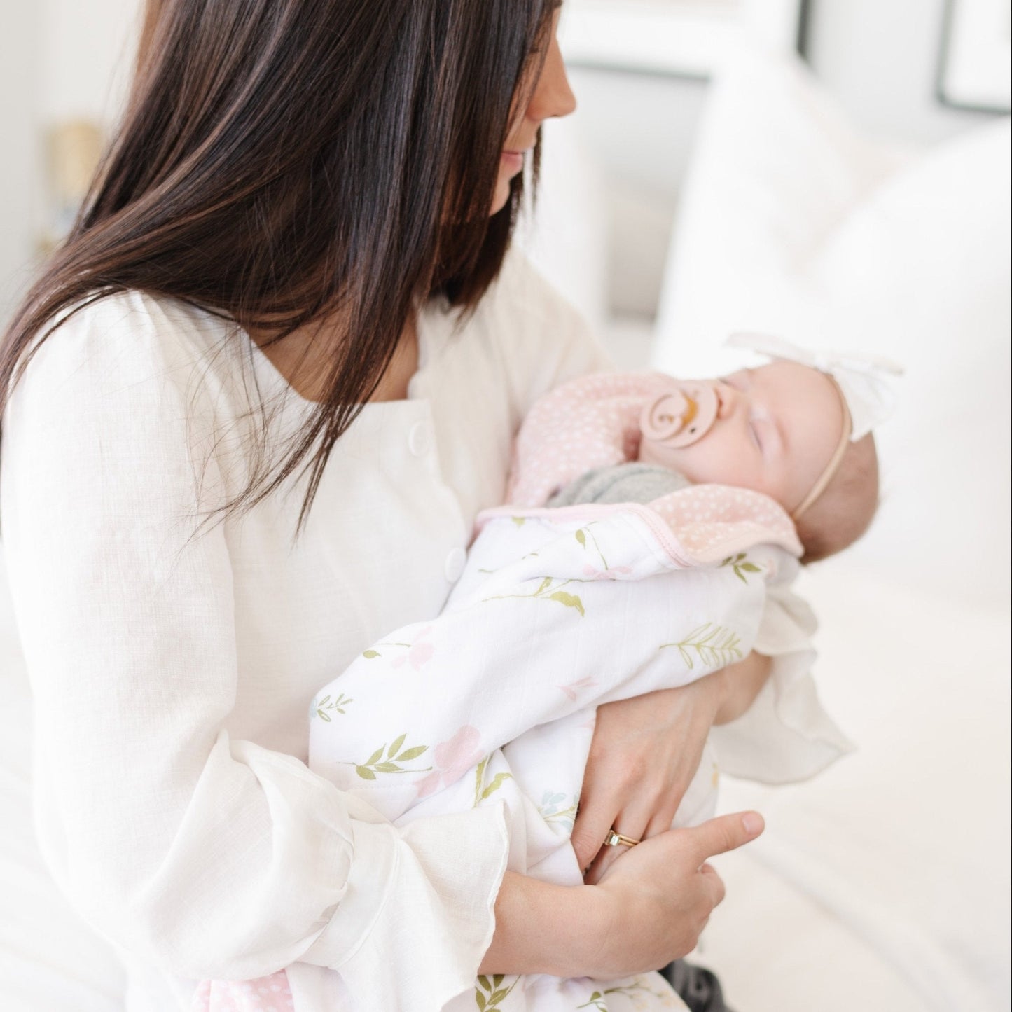 A mother holds her baby girl in a luxuriously soft, reversible muslin quilt. The soft blanket is a luxury blanket and a lightweight and airy baby blanket as well as a Saranoni blanket. One side of the baby quilt features a delicate floral print swaddle design with soft pink trim, while the reverse showcases a blush pink dotted pattern, offering versatility and timeless style. This ultra-soft, baby blanket is perfect for snuggles, tummy time, stroller rides, or nursery decor.