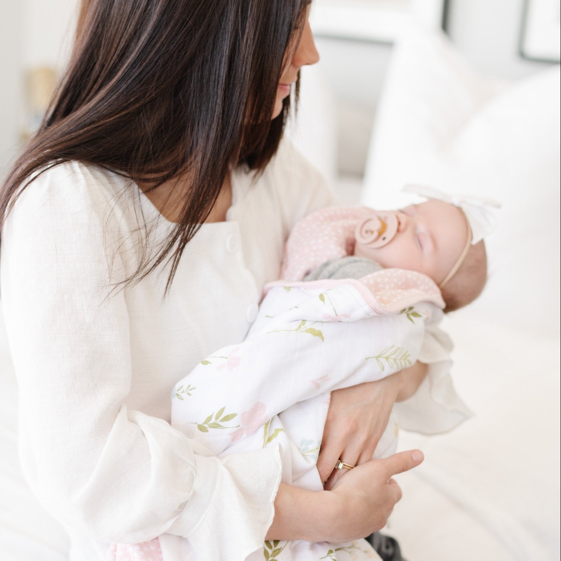 A mother holds her baby girl in a luxuriously soft, reversible muslin quilt. The soft blanket is a luxury blanket and a lightweight and airy baby blanket as well as a Saranoni blanket. One side of the baby quilt features a delicate floral print swaddle design with soft pink trim, while the reverse showcases a blush pink dotted pattern, offering versatility and timeless style. This ultra-soft, baby blanket is perfect for snuggles, tummy time, stroller rides, or nursery decor.