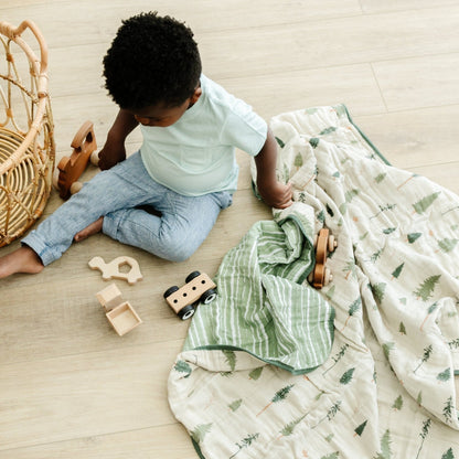 A little boy is playing with toys and is sitting by a cotton muslin quilt that has a pine tree and green striped pattern on it. This soft blanket is a Saranoni blanket and is a great baby blanket or toddler blanket. The adorable pattern makes it a great addition to a nursery or bedroom.