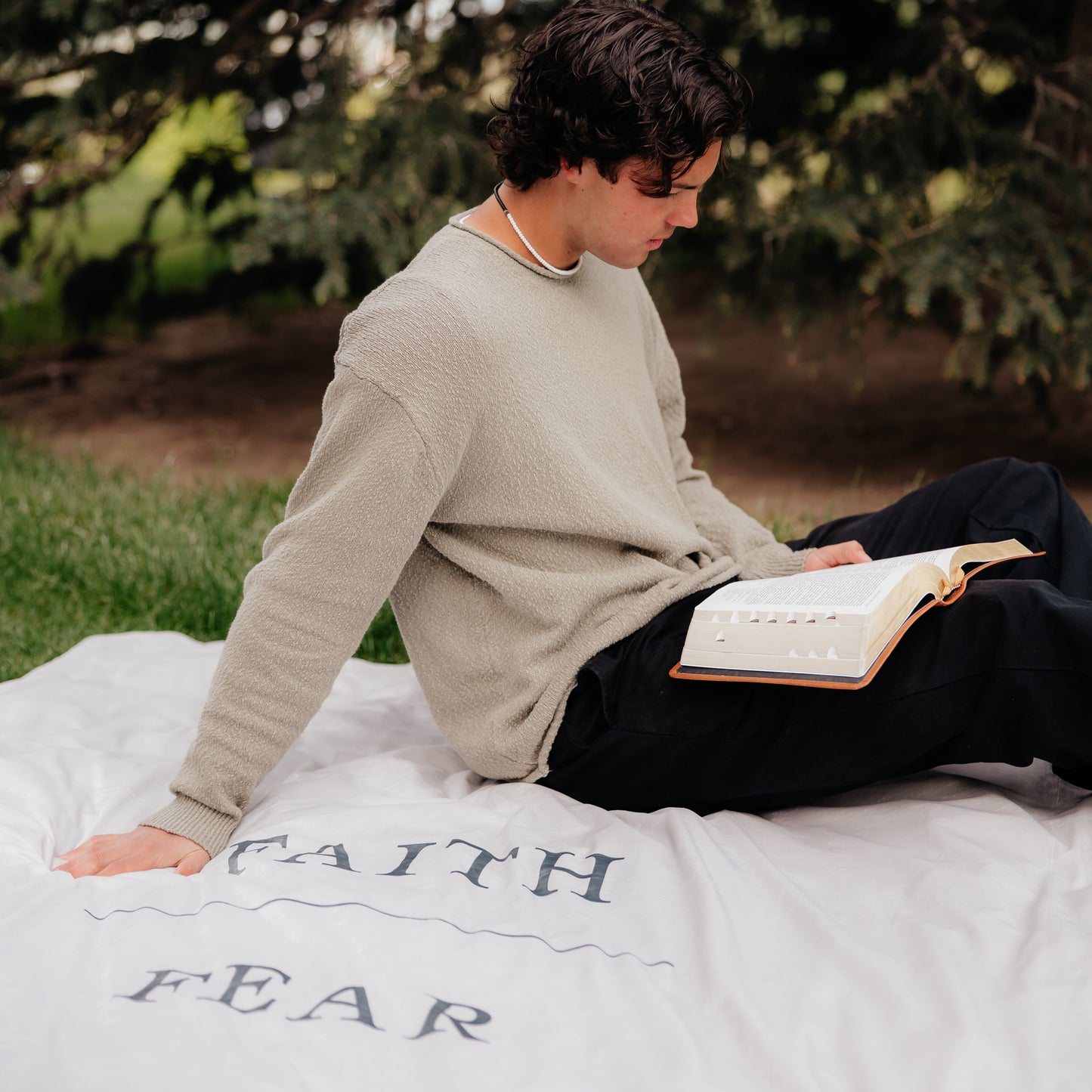 Young man reading scriptures while sitting on a shell cream Christian blanket featuring the words "Faith Over Fear," faith-inspired throw blanket offering comfort, encouragement, and a reminder to trust God through life's challenges.
