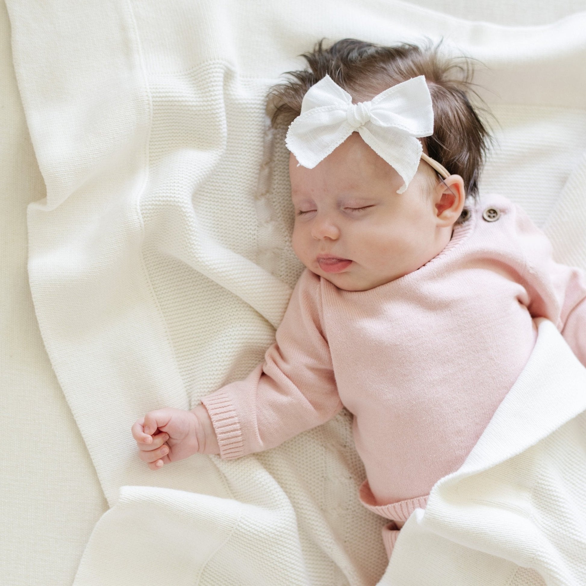 A baby girl lays on a Saranoni Heirloom Knit Blanket. She is wearing a light pink, 100% cotton baby outfit that combines style and comfort. The Saranoni cloud-soft cotton set features a breathable waistband, loosely fitted sleeves, and elevated button details, making it perfect for everyday wear or special occasions. This heirloom-quality baby clothing offers timeless style and ultimate comfort. Ideal for parents searching for luxury baby clothes, soft cotton baby sets, or high-quality infant outfit.