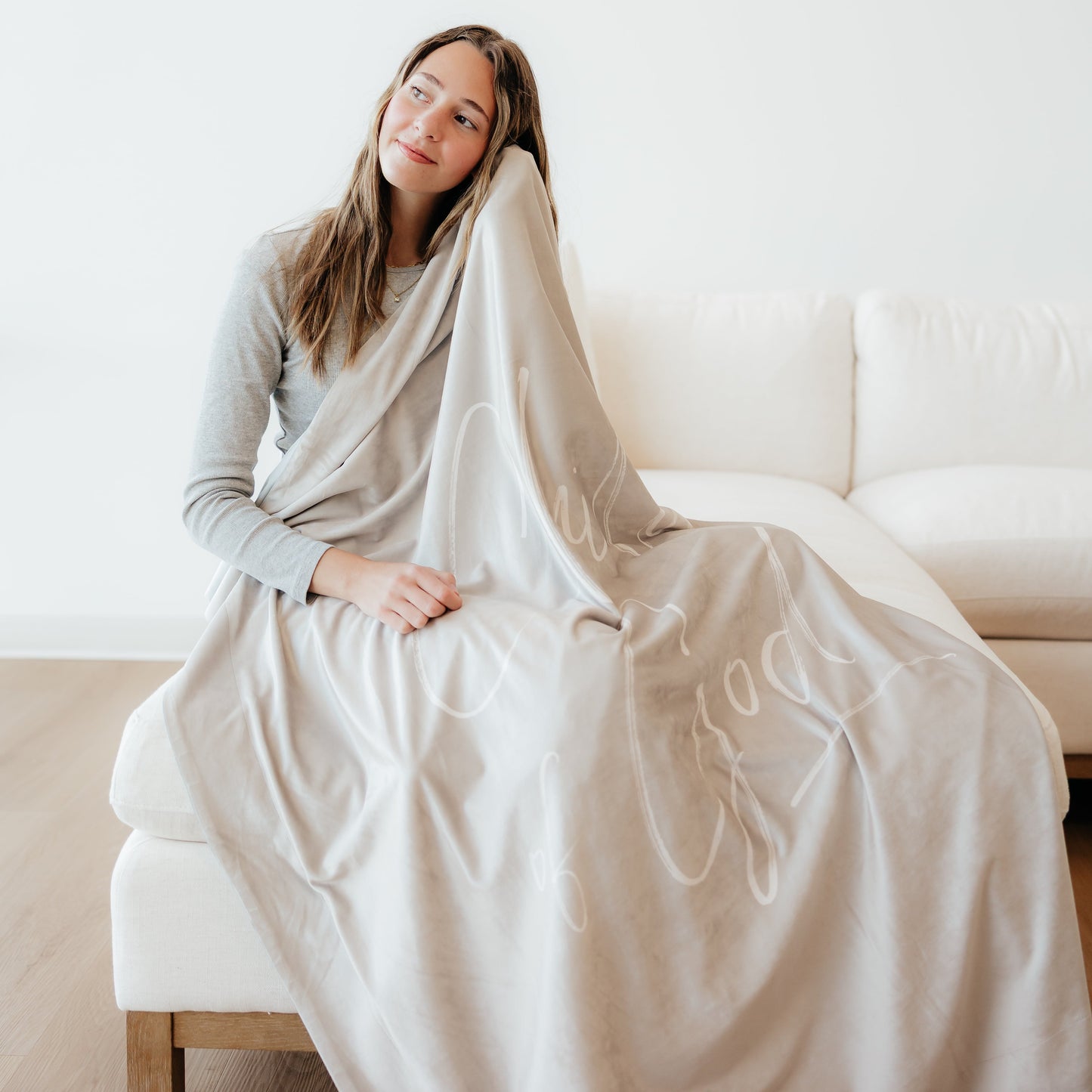 Woman wrapped in a soft cream Christian blanket featuring the words "Child of God" while sitting on a couch in a cozy home setting.