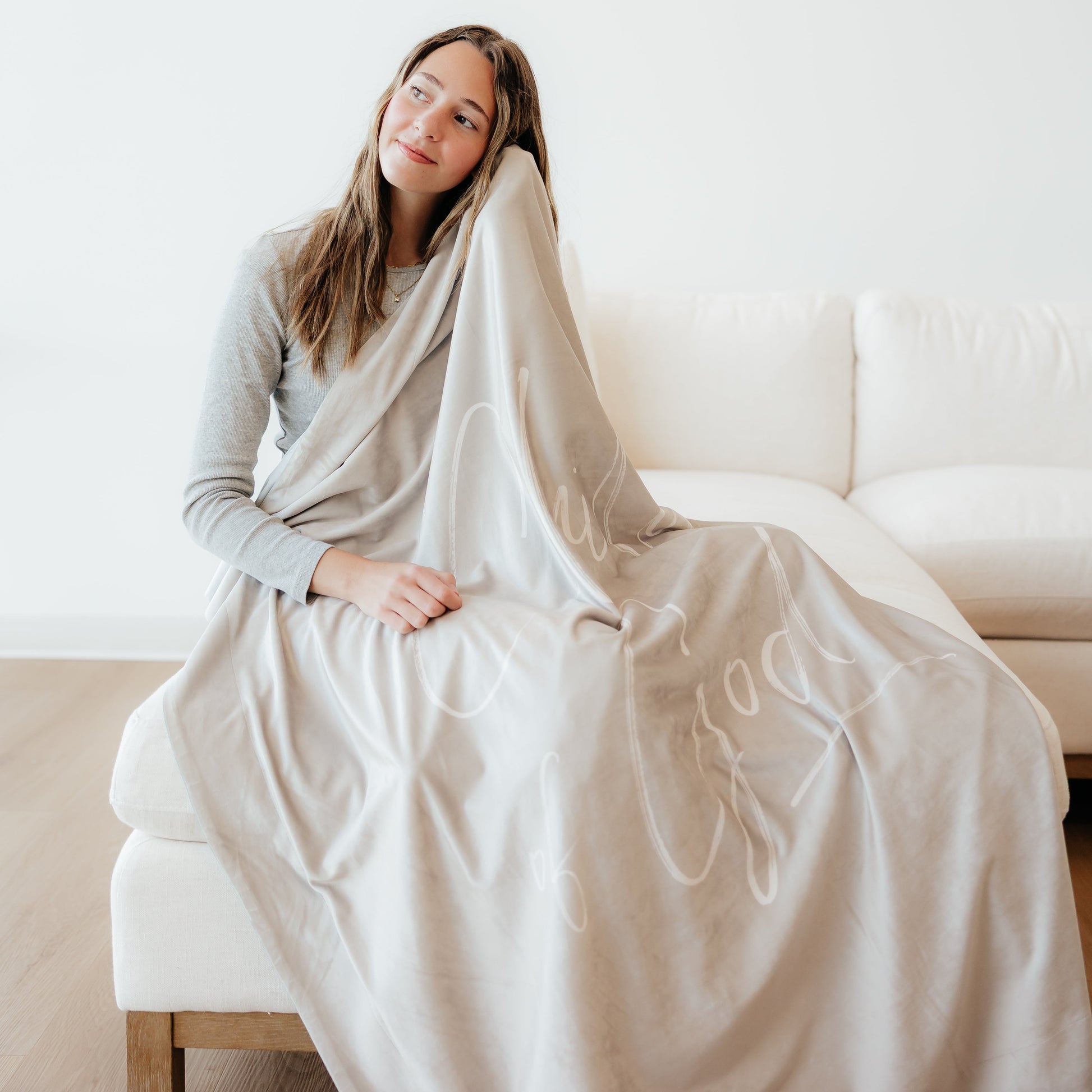 Woman wrapped in a soft cream Christian blanket featuring the words "Child of God" while sitting on a couch in a cozy home setting.