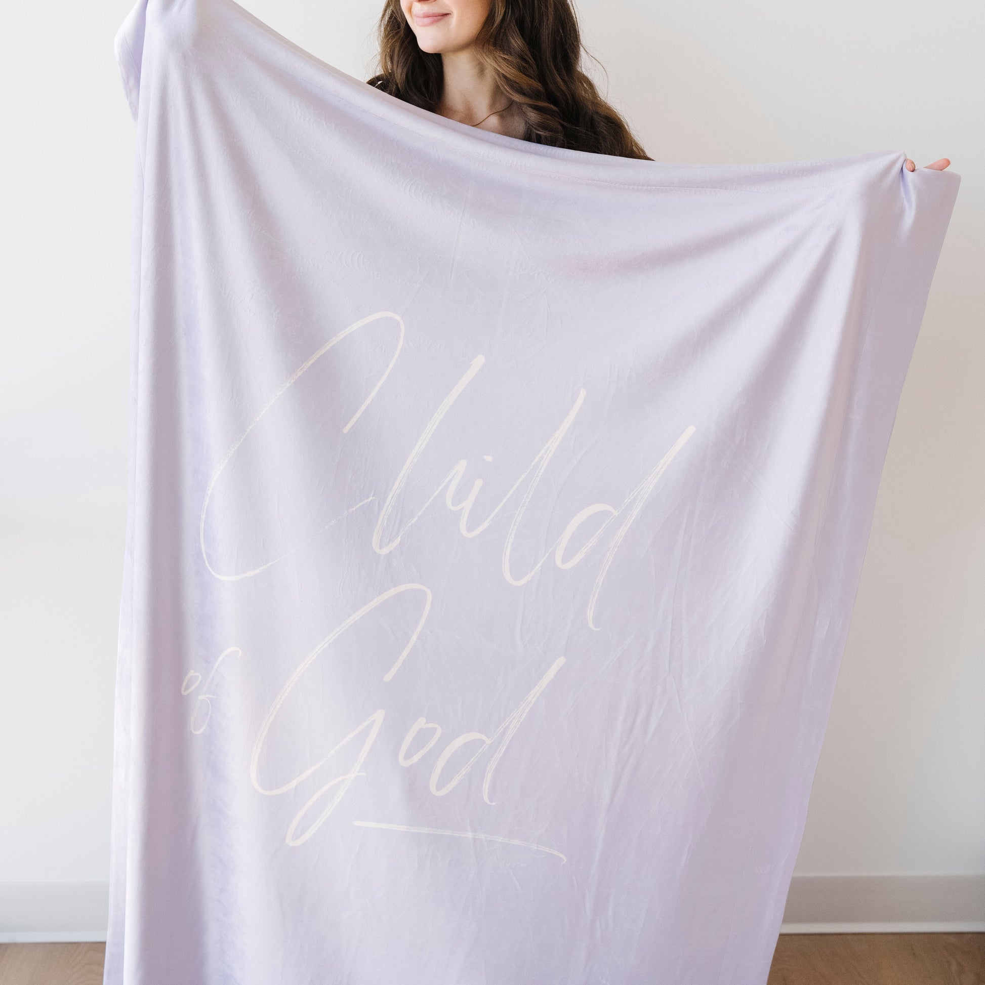 Woman holding a soft lilac Christian blanket featuring the words "His Child" in elegant cursive lettering.