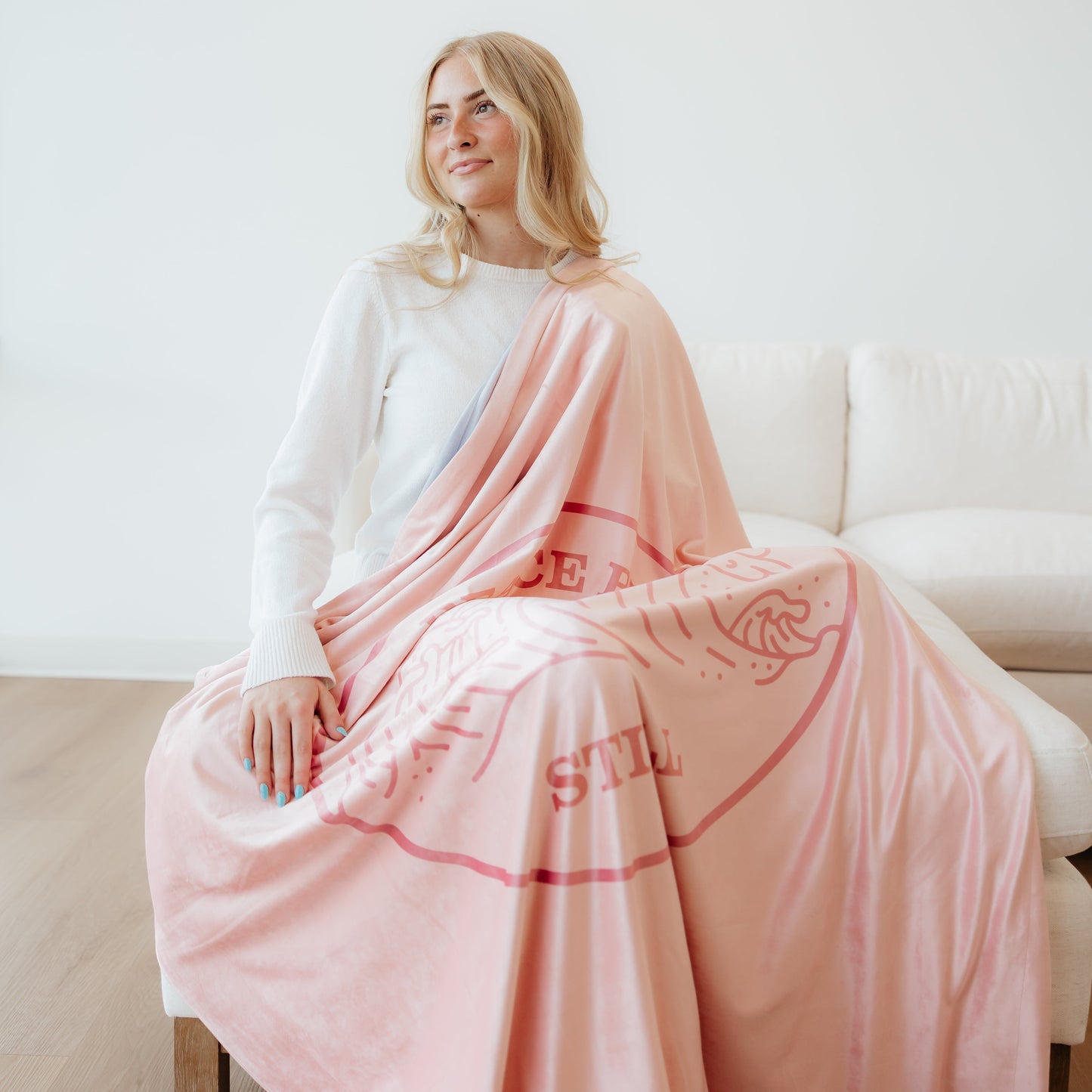 Woman sitting on a couch wrapped in a blush pink Christian blanket featuring the Peace Be Still scripture design and ocean wave artwork.