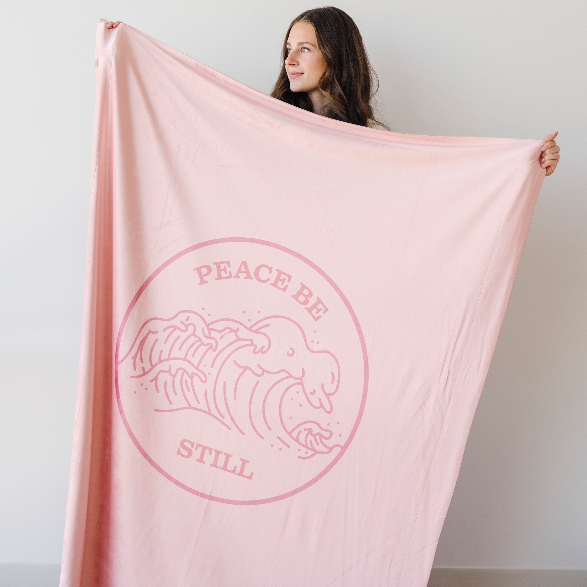 Woman holding a blush pink Christian blanket featuring the Peace Be Still scripture design and ocean wave artwork.