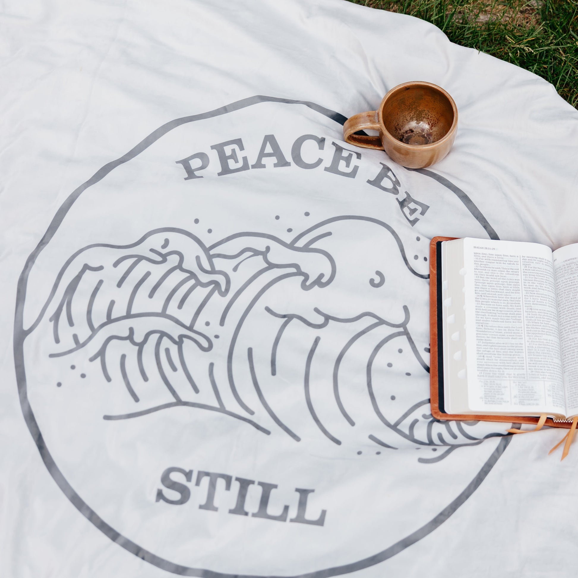 Peace Be Still blanket displayed outdoors with an open Bible and ceramic mug, featuring a calming wave design inspired by scripture.