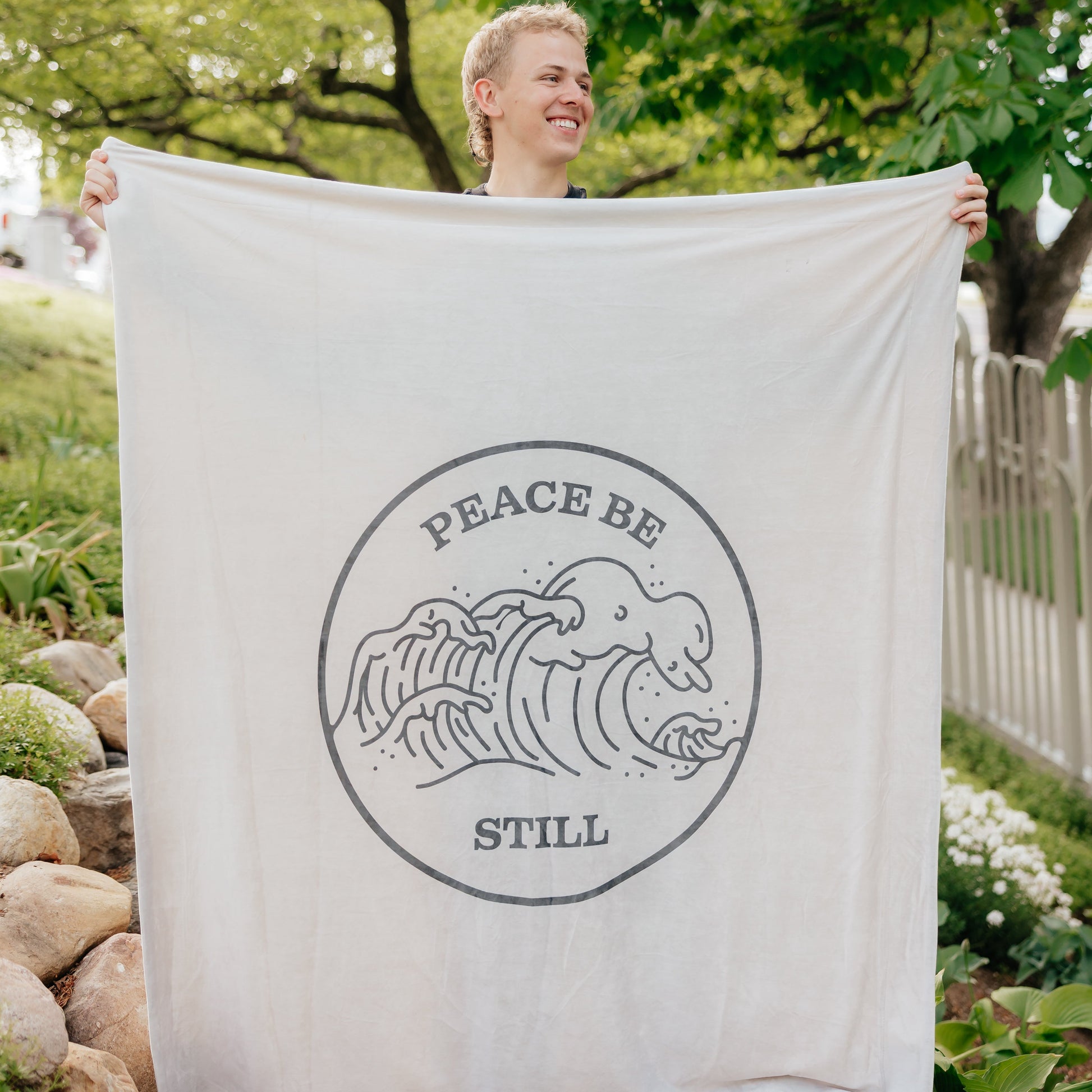 Person holding a Christian blanket featuring the Peace Be Still scripture design and ocean wave artwork outdoors.