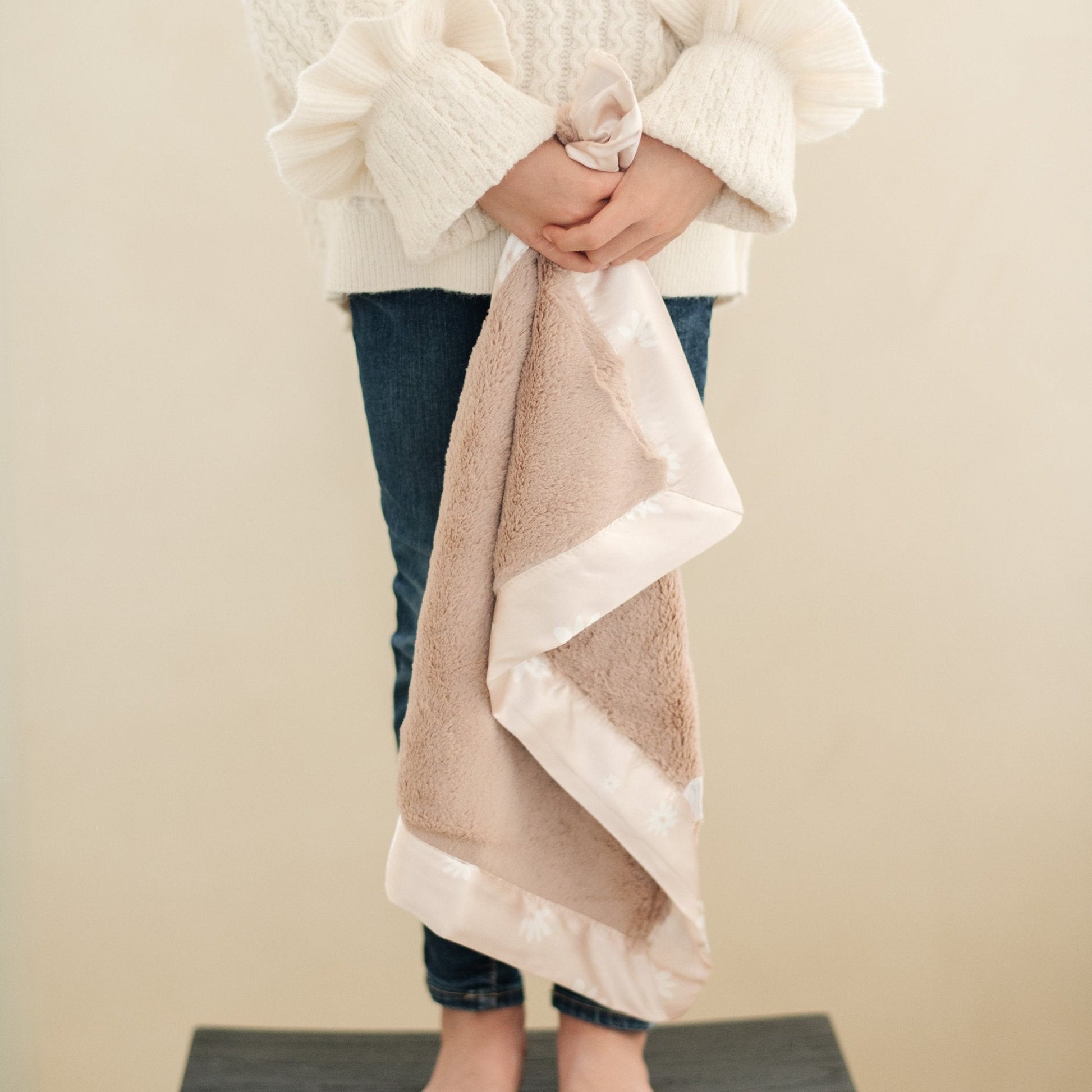 A little girl holds a satin back blanket. The lush blanket is light pink with a light pink satin border that has a daisy design on it. The satin back blanket is a lush blanket with a satin border and back. The soft blanket is a luxury blanket and a Saranoni blanket. This mini blanket is an adorable baby blanket and perfect for littles. 