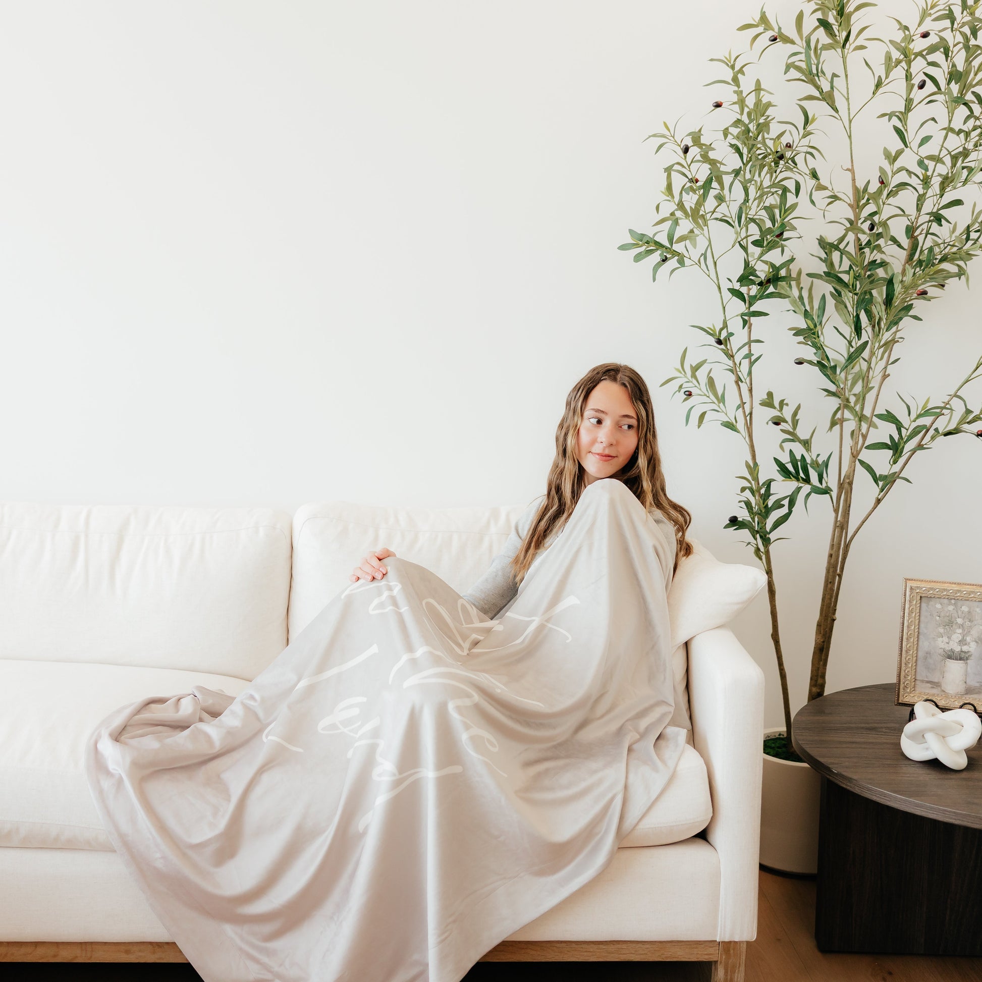 Woman relaxing on a couch wrapped in a light cobblestone gray Christian blanket featuring the words "Trust in the Lord," soft faith-inspired throw blanket for comfort, encouragement, and peace.
