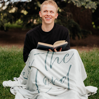 Person reading a Bible outdoors while wrapped in a light rosemary green Christian blanket featuring the words "Trust in the Lord," faith-inspired throw blanket for comfort, encouragement, and reflection.
